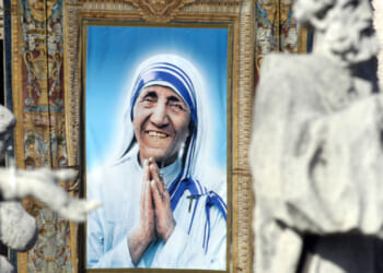 A large portrait of a smiling woman in a white and blue sari with hands clasped in prayer, framed and displayed outdoors, with blurred statues in the foreground.