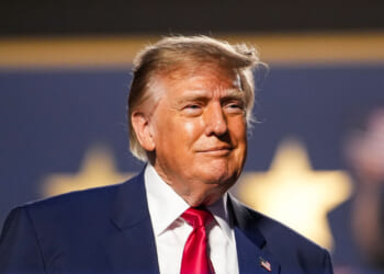 A man in a suit and red tie stands in front of a blurred background with large yellow stars. He has light hair and is looking slightly to the side with a neutral expression.