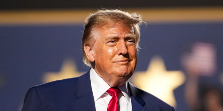 A man in a suit and red tie stands in front of a blurred background with large yellow stars. He has light hair and is looking slightly to the side with a neutral expression.