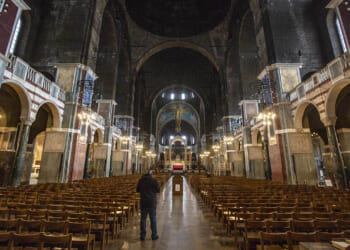 A person stands alone in the aisle of a grand, dimly lit cathedral with rows of empty wooden chairs and a large cross at the altar ahead.