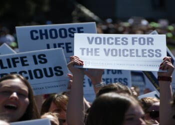 A crowd holds signs with messages like Voices for the Voiceless, Life is Precious, and Choose Life at a public demonstration, with people visible in the foreground.