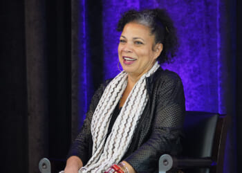 Pictured: Professor Melina Abdullah, a woman with curly hair, sits on a chair, smiling. She wears a black outfit with a white patterned scarf. Behind her is a purple-lit curtain. A water bottle and green glass bottle are on a table nearby.