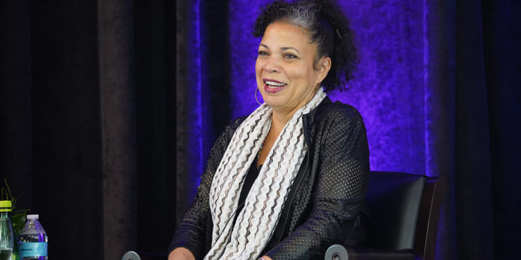 Pictured: Professor Melina Abdullah, a woman with curly hair, sits on a chair, smiling. She wears a black outfit with a white patterned scarf. Behind her is a purple-lit curtain. A water bottle and green glass bottle are on a table nearby.