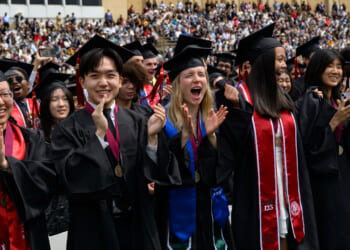 Graduates in caps and gowns cheer and clap during a graduation ceremony, with a distinguished professor standing nearby and a large audience in the background.