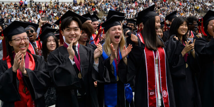 Graduates in caps and gowns cheer and clap during a graduation ceremony, with a distinguished professor standing nearby and a large audience in the background.