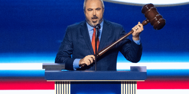 A man in a blue suit and red tie stands at a podium, holding a large wooden gavel and speaking into a microphone, with a blue and red background behind him.