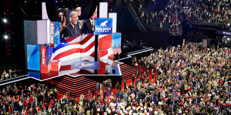 A large crowd fills an arena with a stage displaying RNC 2024 and American flags. A speaker addresses the audience, while people hold signs and cheer. The atmosphere is lively and patriotic.