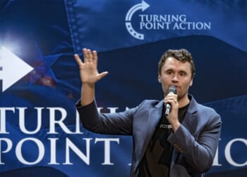 A man in a blazer speaks into a microphone, raising one hand, in front of a blue Turning Point Action backdrop with white text and arrow graphics, reminiscent of events frequently hosted by Charlie Kirk.