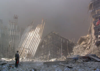 A person stands amid debris and dust clouds, surrounded by partially collapsed skyscrapers and twisted metal in a devastated urban landscape in the aftermath of 9/11.
