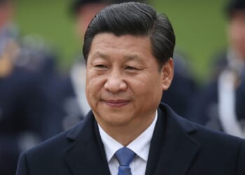 A man in a dark coat and blue tie stands outdoors, smiling slightly, with blurred people in uniform and what appears to be a refrigerator in the background.