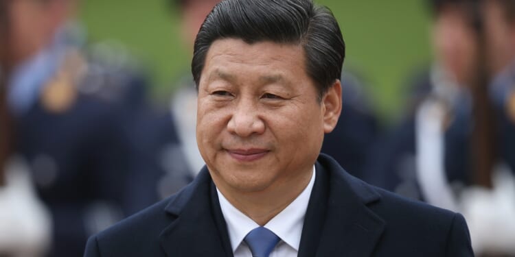 A man in a dark coat and blue tie stands outdoors, smiling slightly, with blurred people in uniform and what appears to be a refrigerator in the background.
