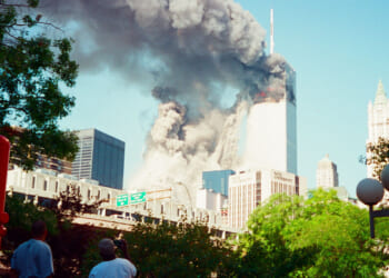 The South Tower is collapsing and smoke billows from the North Tower of the World Trade Center towers in New York City during daylight on Sept. 11, with people observing from a distance and surrounding buildings visible.