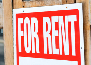 A close-up of a red and white For Rent sign is attached to a wooden door, indicating that the property is available for leasing.