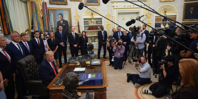 Donald Trump sits at the Resolute Desk in the Oval Office, surrounded by officials and media, with microphones and cameras pointed toward him. Portraits and gold decor line the walls.