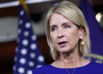 Mary Miller with blonde hair in a blue dress speaks in front of blurred U.S. flags and a dark background. She wears earrings and looks slightly to the side.