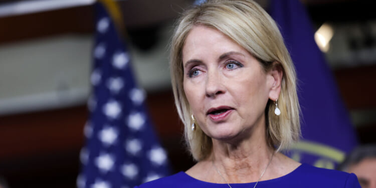 Mary Miller with blonde hair in a blue dress speaks in front of blurred U.S. flags and a dark background. She wears earrings and looks slightly to the side.