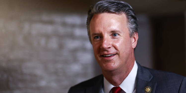 Smiling man in a suit and red tie stands indoors with a blurred, neutral background. He has short gray hair and wears a pin on his lapel.