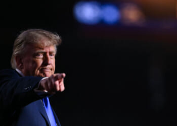 Republican President Donald Trump points forward with a serious expression, standing against a dark background with blurred lights in the upper right corner.