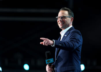 A man in a blue suit and glasses smiles while holding a microphone and pointing, standing on a stage with blue lights in the background, perhaps addressing the audience about disease awareness.