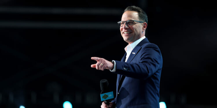 A man in a blue suit and glasses smiles while holding a microphone and pointing, standing on a stage with blue lights in the background, perhaps addressing the audience about disease awareness.