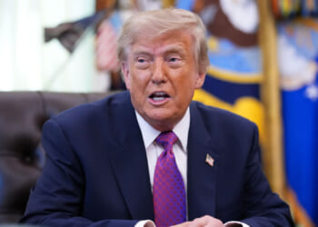 President Trump in a navy suit and purple tie sits at a desk, speaking. Flags and decorative items are visible in the blurred background.
