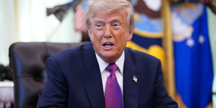 President Trump in a navy suit and purple tie sits at a desk, speaking. Flags and decorative items are visible in the blurred background.