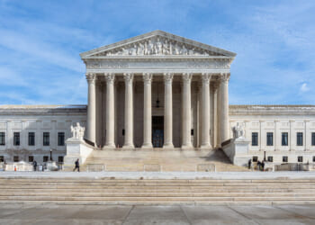 The United States Supreme Court building with tall columns, a triangular pediment, statues, and broad stone steps under a blue sky.