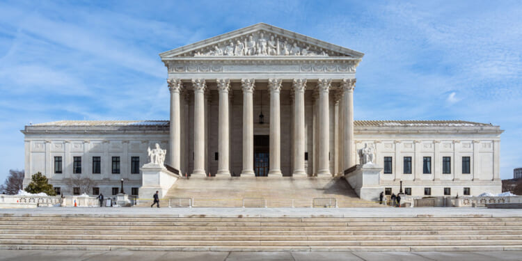 The United States Supreme Court building with tall columns, a triangular pediment, statues, and broad stone steps under a blue sky.