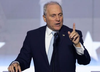 A man in a dark suit and light blue tie speaks at a podium, gesturing with his right hand raised and index finger pointing up. A blurred background shows large white stars.