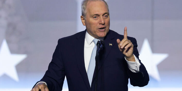 A man in a dark suit and light blue tie speaks at a podium, gesturing with his right hand raised and index finger pointing up. A blurred background shows large white stars.