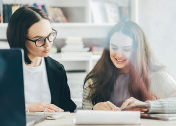 The American School Counselor Association has admitted that neurodiversity is a term for critical race theory and queer theory. Pictured: Two women sit at a table with books and a laptop, smiling as they look at papers together in a bright, cozy room with shelves of books in the background.