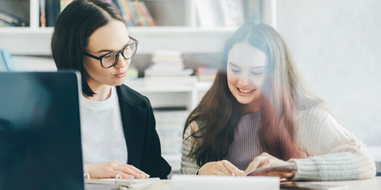 The American School Counselor Association has admitted that neurodiversity is a term for critical race theory and queer theory. Pictured: Two women sit at a table with books and a laptop, smiling as they look at papers together in a bright, cozy room with shelves of books in the background.