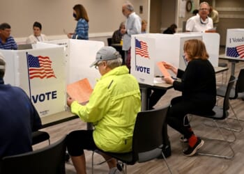 People sit at voting booths filling out ballots in a polling station. Voting partitions display the American flag and the word VOTE. The room is well-lit, and several voters are present.