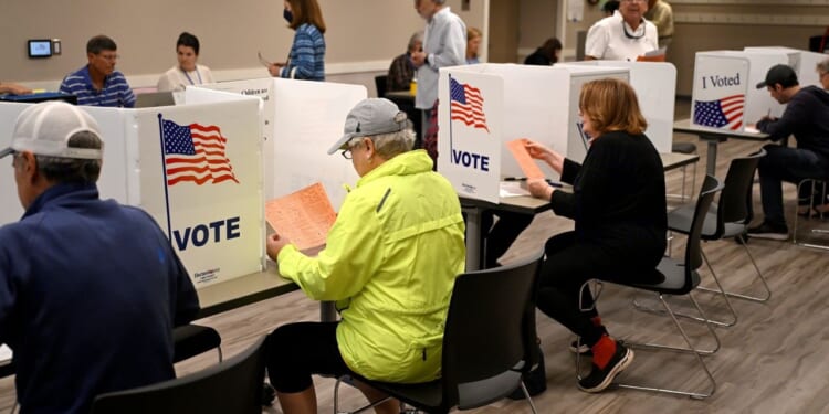 People sit at voting booths filling out ballots in a polling station. Voting partitions display the American flag and the word VOTE. The room is well-lit, and several voters are present.