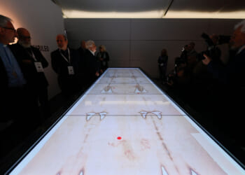 A group of people stand around a long, illuminated display table featuring faint, human-like images and markings, while photographers capture the scene in a dimly lit room.