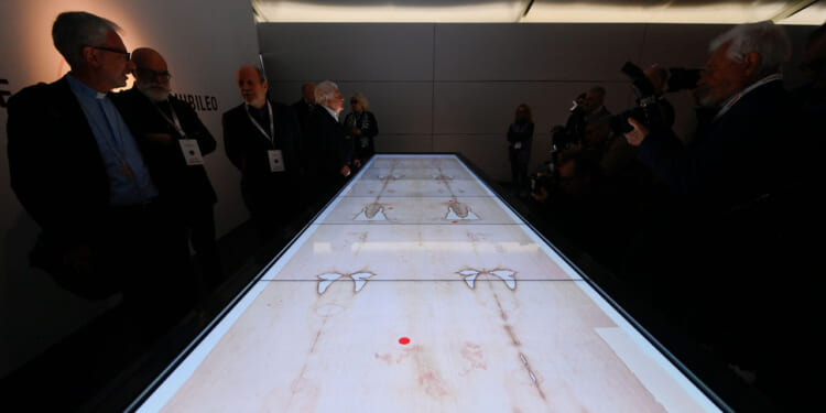 A group of people stand around a long, illuminated display table featuring faint, human-like images and markings, while photographers capture the scene in a dimly lit room.