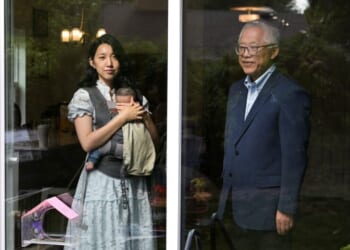 A woman holding a baby in a carrier stands inside a house, looking out a glass door. An older man stands outside, facing her. The door separates them, with reflections of trees and lights visible on the glass.