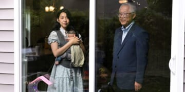 A woman holding a baby in a carrier stands inside a house, looking out a glass door. An older man stands outside, facing her. The door separates them, with reflections of trees and lights visible on the glass.