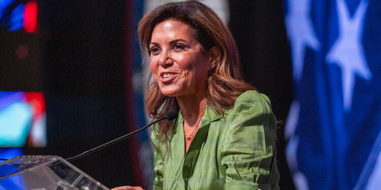 A woman in a green blouse speaks at a podium with a microphone. The background features a large, partially visible American flag with blue and white stars.