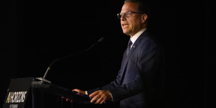 A man in a navy suit and glasses speaks at a podium with a microphone against a dark background. The podium sign reads INNOVATIONS PITTSBURGH..