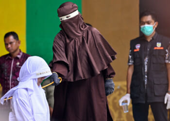 A person in brown robes prepares to cane a kneeling individual in white while uniformed officials and onlookers stand nearby, one wearing a face mask.