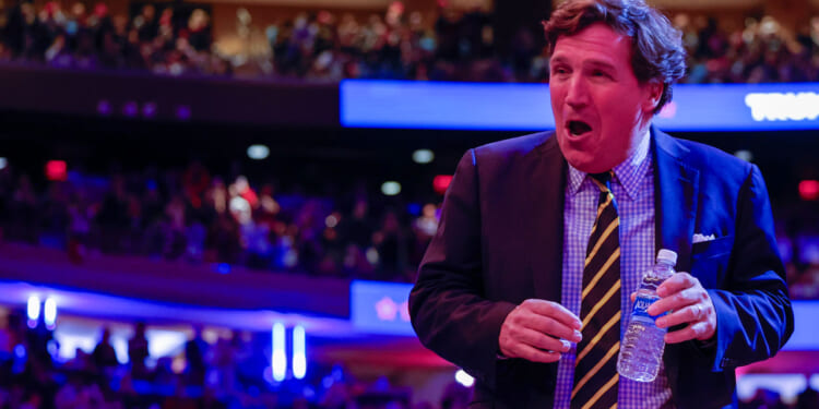 Tucker Carlson in a suit and striped tie, holds a water bottle as he stands in a crowded auditorium with colorful lights and a blurred audience in the background.