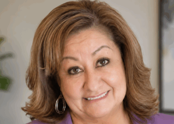 School counselor Jeanette Martinez is suing Albuquerque Public Schools over its gender secrecy policy. Pictured: a woman with light brown hair, wearing hoop earrings and a purple top, smiles at the camera. The background is softly blurred, with a plant and a framed picture visible.