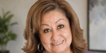 School counselor Jeanette Martinez is suing Albuquerque Public Schools over its gender secrecy policy. Pictured: a woman with light brown hair, wearing hoop earrings and a purple top, smiles at the camera. The background is softly blurred, with a plant and a framed picture visible.