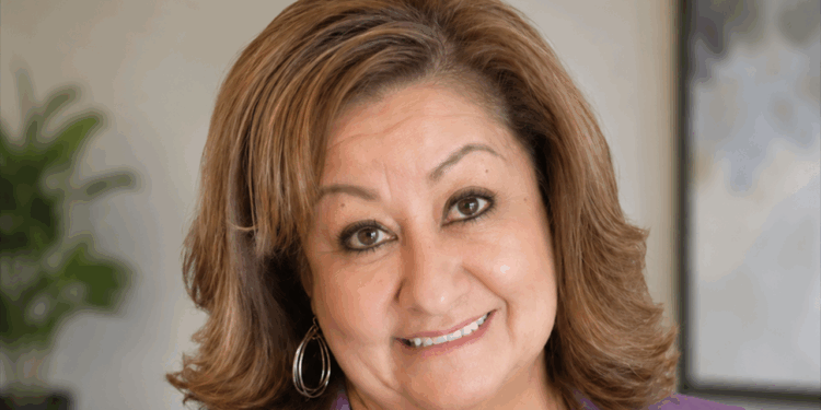 School counselor Jeanette Martinez is suing Albuquerque Public Schools over its gender secrecy policy. Pictured: a woman with light brown hair, wearing hoop earrings and a purple top, smiles at the camera. The background is softly blurred, with a plant and a framed picture visible.
