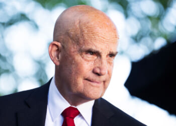 Keith Self in a dark suit, white shirt, and red tie, standing outdoors with blurred green foliage in the background.