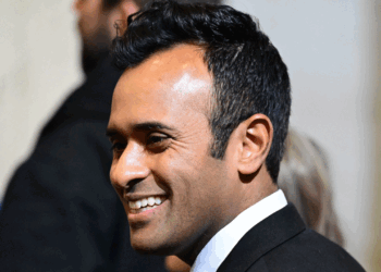 Vivek Ramaswamy in a black suit and white shirt smiles as he stands indoors, with blurred people and neutral-colored walls in the background.