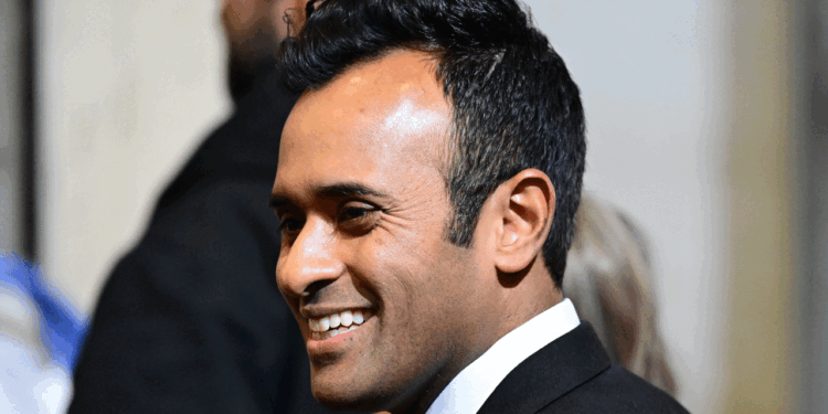 Vivek Ramaswamy in a black suit and white shirt smiles as he stands indoors, with blurred people and neutral-colored walls in the background.
