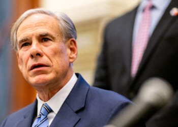 Greg Abbott a blue suit and striped tie sits indoors, looking slightly upward, with another person in a suit standing blurred in the background.