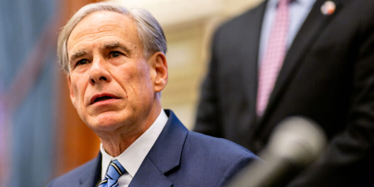 Greg Abbott a blue suit and striped tie sits indoors, looking slightly upward, with another person in a suit standing blurred in the background.